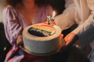 привітання з днем народження свекрусі — A vibrant birthday cake featuring a rainbow design and burning candle held by two people. | ITmaxService