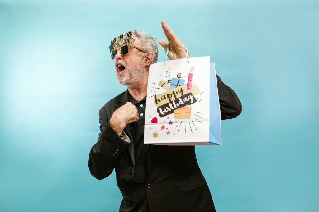привітання з днем народження дружини — Senior man wearing sunglasses, holding a birthday gift bag on a light blue background. | ITmaxService